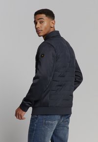 Marineblauwe pufferjack met een geborduurde textuur op de achterkant, een gladde voorkant, ribbels aan de manchetten en kraag. Accentueert met een hexagonale logo op de schouder.