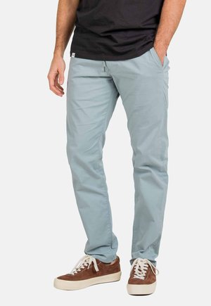 Light blue cotton trousers with a straight fit, featuring side pockets and a zipper. Paired with brown sneakers and a black shirt.