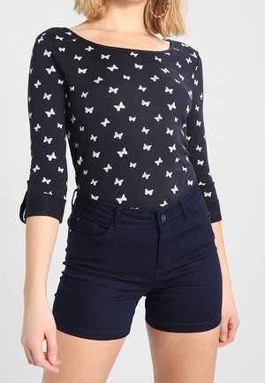 Kobieta ubrana w czarną bluzkę z długim rękawem z białym nadrukiem motyli i dopasowane granatowe szorty stoi na jednolitym tle.