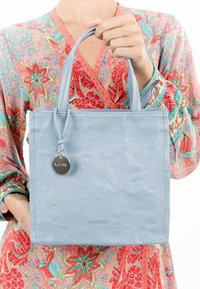 Light blue leather tote bag with a structured shape, featuring two handles and a round metal tag. Embossed logo on the front.
