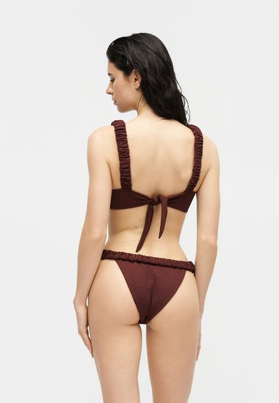 Femme aux cheveux foncés portant un bikini marron foncé texturé avec des bretelles froncées, vue de dos sur un fond clair uni.