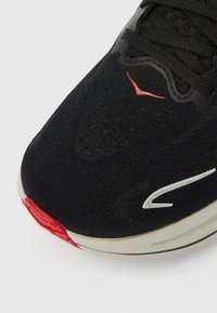 Μαύρο αθλητικό παπούτσι με υφή στο πάνω μέρος, με κόκκινη λεπτομέρεια στη μύτ η και λευκό λογότυπο στη πλευρά. Ελαφριά κατασκευή.