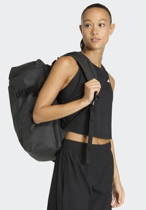 Mochila negra con correas ajustables, lado texturizado y múltiples compartimentos. Combinada con una camiseta corta negra y pantalones holgados.