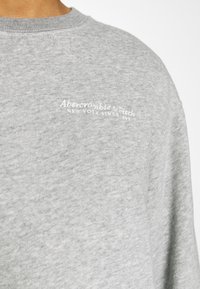 Harmaa puuvillainen collegepaita, jossa on pyöreä pääntie, resorit hihansuissa ja pieni valkoinen kirjailtu logo rinnassa. Kankaan pinta on teksturoitu.