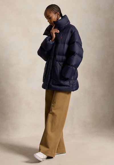 Femme aux cheveux tressés portant une doudoune bleu marine, un pantalon large couleur beige et des baskets blanches, debout devant un fond neutre.