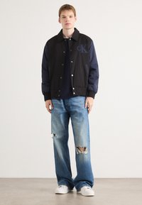 Black and navy bomber jacket with a CK logo, paired with light blue distressed wide-leg jeans and white sneakers. Smooth fabric texture.