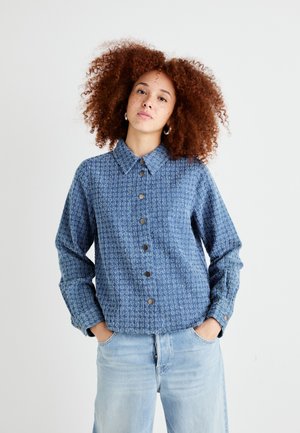 Blue button-up shirt with a textured pattern, featuring a collar and long sleeves. It has dark buttons and is paired with light blue jeans.