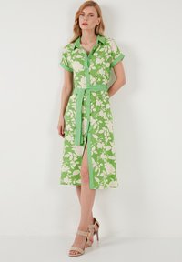 Grün gemustertes Hemdkleid mit kurzen Ärmeln, Knopfleiste, gebundenem Taillengurt und knielanger Länge. Mit weißen Blumenmustern und einem seitlichen Schlitz.