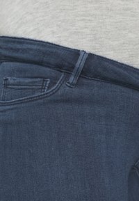 Primer plano de unos vaqueros de mezclilla azul oscuro con el bolsillo delantero, el lazo del cinturón y la cinturilla de tela gris visibles.