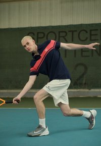 Tenisový hráč v námorníckej a ružovej polokošeli, bielych šortkách a šedých športových topánkach s ružovými akcentmi, pripravený udeliť úder lopte na modrom ihrisku.