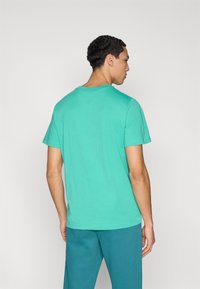 Πράσινη μπλούζα με κοντά μανίκια και στρογγυλή λαιμόκοψη, κατασκευασμένη από μαλακό ύφασμα, συνδυασμένη με μπλε-πράσινο παντελόνι, σε προβολή από πίσω.