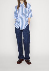 Camicia a righe blu e bianca, oversize, con maniche corte e taschino sul petto, abbinata a pantaloni cargo blu navy e scarpe marroni.