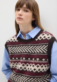 Strickweste mit geometrischem Muster in Burgunderrot, Creme und Braun, getragen über einem hellblauen Hemd mit Kragen. Texturierte Wollmischung.
