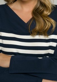 Donna con lunghi capelli mossi, che indossa una maglietta blu navy con righe orizzontali bianche sul petto, con le braccia incrociate.