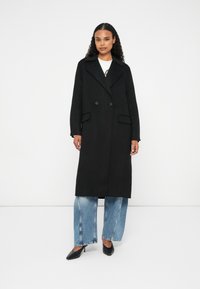 Mujer vestida con un largo abrigo negro de doble botonadura sobre jeans y zapatos de tacón negros, de pie contra un fondo blanco liso.