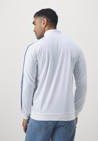 Hvit langermet jakke med høy krage, med blå striper langs skuldrene og mykt, teksturert stoff.