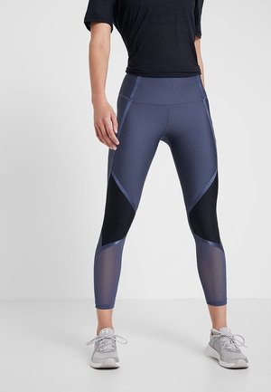 Personne portant des leggings bleu marine taille haute avec des panneaux noirs et en mesh, associés à des chaussures de sport grises et un haut noir à manches courtes.