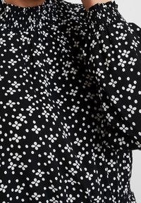 Svart blus med vitt blommönster; har samlad halsringning och puffärmar; mjukt, texturerat tyg med jämnt fördelade mönster.