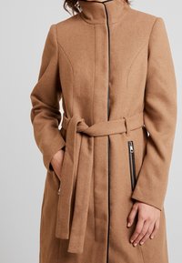 Vaalea villasekoitteen takki, jossa on korkea kaulus, istuva siluetti, vyöllä varustettu vyötärö ja vetoketjulliset sivutaskut. Kontrastiväriset tikkaukset vetoketjun ympärillä.