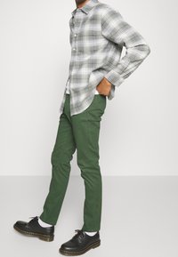 Mężczyzna w zielonych obcisłych spodniach, czarnych skórzanych butach, białych skarpetkach i szaro-białej kraciastej koszuli z długim rękawem, stojący z jedną ręką w kieszeni.