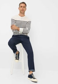 Maglione a righe bianche e blu navy, pantaloni in denim scuro e sneakers blu navy con suole crema. Modella seduta su uno sgabello bianco contro uno sfondo neutro.