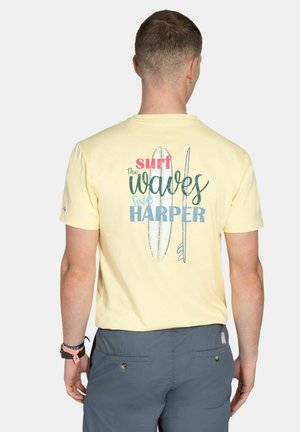 Hombre que lleva una camiseta de color amarillo pálido con tablas de surf y el texto "surf the waves live HARPER" impreso en la parte trasera, combinado con pantalones cortos azules.