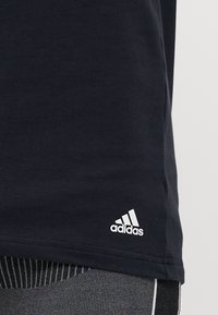 Schwarzes Baumwoll-T-Shirt mit abgerundetem Saum, versehen mit einem kleinen weißen Adidas-Logo unten links, kombiniert mit strukturierten grauen Leggings.