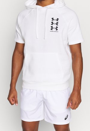 Mann trägt einen weißen Kurzarm-Hoodie mit Under Armour-Logo und weiße sportliche Shorts mit Asics-Logo, steht vor einem einfarbigen Hintergrund.