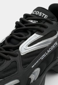 Baskets noires Lacoste avec des détails argentés, des lacets noirs et le texte "From the court to the streets since 1933" sur le côté.