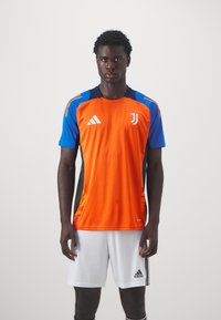 Camisa de futebol da Juventus com corpo laranja, mangas azuis e laterais pretas. Apresenta o logotipo da Adidas e um padrão de gradiente texturizado perto da bainha.