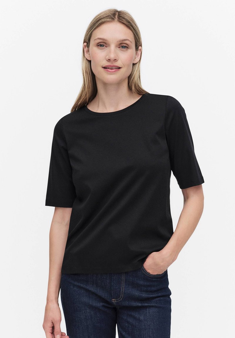 Kvinde med langt blondt hår iført en sort top med korte ærmer og mørkeblå jeans, stående med den ene hånd i lommen mod en hvid baggrund.