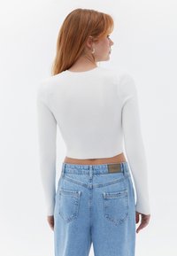 Weißes geripptes Langarm-Crop-Top kombiniert mit hoch geschnittenen hellblauen Jeans. Auffällige Details sind sichtbare Nähte und klassische Taschen.