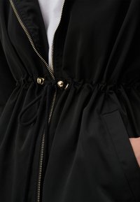 Zwarte jas met een tunnelkoord in de taille, gouden ritssluiting en zakken. Het materiaal lijkt glad met een subtiele glans.