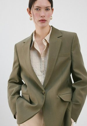 Mujer con blazer verde oliva sobre camisa beige a cuadros, con pendientes de aro dorados, manos en los bolsillos, mirando hacia adelante.