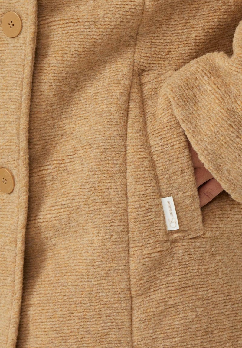 Beige texturierten Stoffmantel mit Knopfverschluss, Seitentasche und einem Markenetikett, der ein weiches, warmes Aussehen präsentiert.