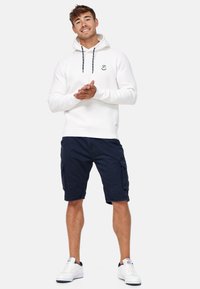 Hoodie blanc avec cordon de serrage, shorts cargo navy et baskets blanches. Le hoodie arbore un logo ainsi que des poignets et un ourlet côtelés.