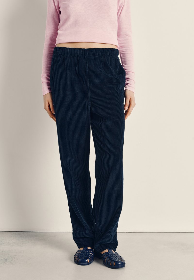 Asmuo dėvi tamsiai mėlynas korduroko kelnes su elastine juosmens dalimi, rožinį ilgomis rankovėmis marškinėlį ir tamsiai mėlynos spalvos pintus slip-on batus, stovintis priešais lygų sieną.