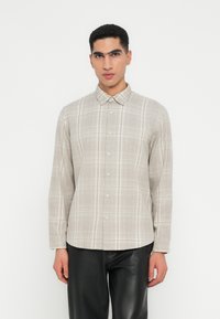 Hombre vestido con una camisa de cuadros beige de manga larga y pantalones de cuero negros, de pie frente a un fondo blanco.