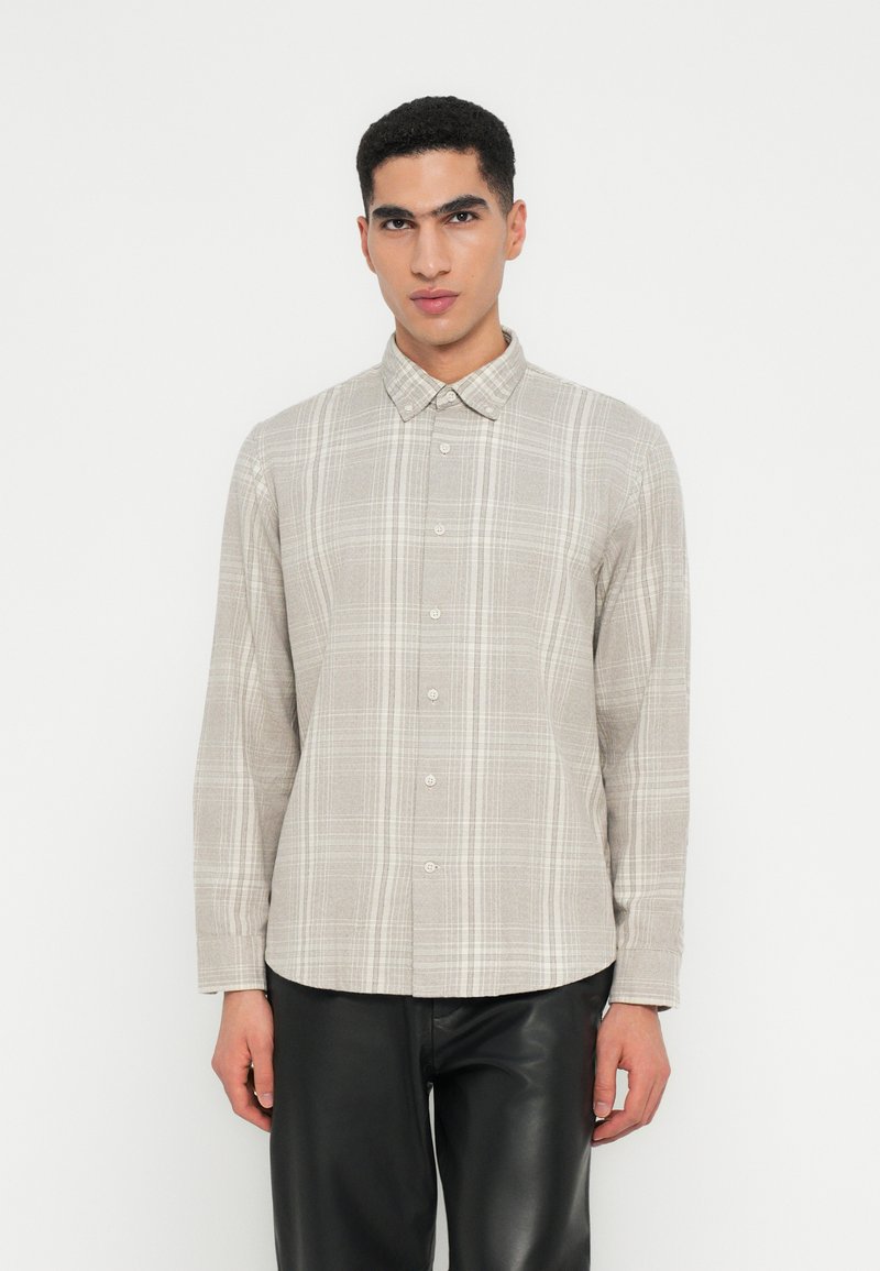 Hombre vestido con una camisa de cuadros beige de manga larga y pantalones de cuero negros, de pie frente a un fondo blanco.