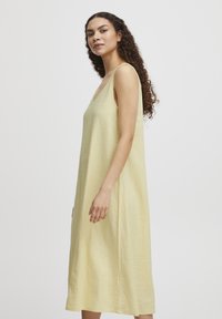 Rochie maxi fără mâneci, de culoare galben deschis, cu o croială relaxată și decolteu rotund, fabricată dintr-un material texturat. Lungime până la jumătatea vițelului.