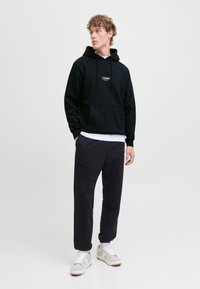 Zwarte hoodie met een voorzak en logo, gecombineerd met donkere broeken en lichtgekleurde sneakers, tegen een effen achtergrond.