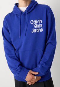 Blauwe katoenen hoodie met trekkoord, voorkeurszak en geborduurd "Calvin Klein Jeans" logo. Miniatuur logo op de linkermouw.