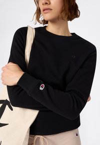 Sudadera negra con cuello redondo y puños de canalé, que presenta un pequeño logo bordado en blanco y rojo en la manga. Material de algodón.