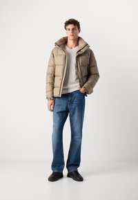 Veste matelassée beige avec fermeture éclair et bouton, dotée d'un col beige. Portée sur une chemise blanche et un jean bleu, complétée par des baskets noires.