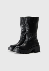 Bottes noires en cuir jusqu'au genou avec un talon épais et une semelle en caoutchouc texturée, présentant des coutures latérales et des languettes pour un enfiler facile.