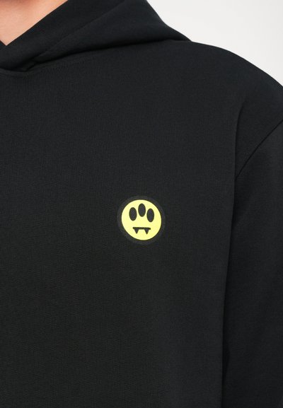 Felpa nera con cappuccio caratterizzata da un piccolo logo tondo giallo con un volto sorridente e zanne, realizzata in tessuto con una finitura testurizzata.
