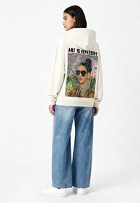 Felpa con cappuccio color crema con una grande stampa sul retro di una donna con occhiali da sole, circondata da elementi tropicali. Il testo dice "L'ARTE È COSTOSA." Indossata con jeans blu larghi.