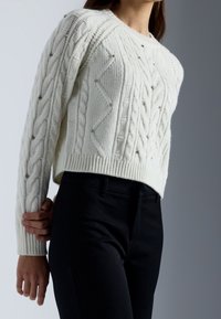 Weißer Pullover aus Kabelstrick mit rundem Ausschnitt, der erhabene Muster und kleine dekorative Verzierungen aufweist, kombiniert mit schwarzen Hosen.