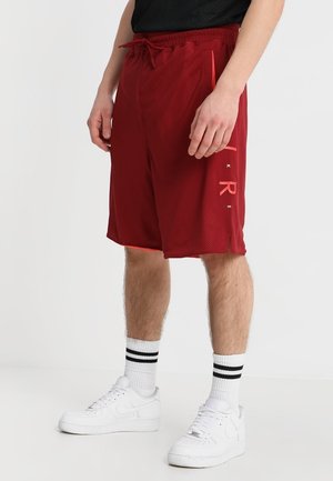 Mężczyzna noszący czerwone sportowe szorty z siateczki ze sznurkiem, białe skarpetki z czarnymi paskami oraz białe trampki, stojący na jednolitym tle.
