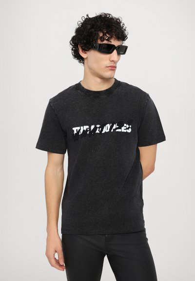 Zwarte katoenen T-shirt met korte mouwen, voorzien van een vervormd wit logo ontwerp over de borst, gecombineerd met zwarte zonnebril.